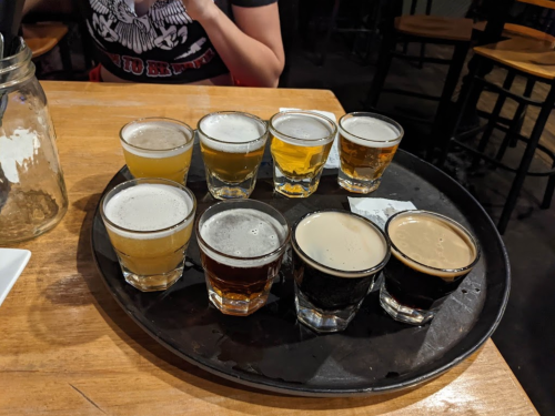 A tray with eight small glasses of different beers, arranged in two rows on a wooden table.