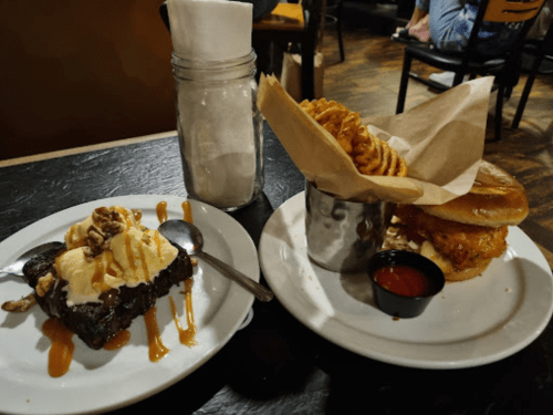 A plate with a brownie topped with ice cream and caramel, and a sandwich with curly fries in a cup on the side.