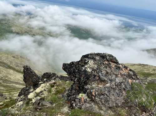 A rocky outcrop on a hillside with clouds rolling over a lush green landscape and a distant ocean view.