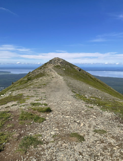 A rocky mountain trail leads to a peak under a clear blue sky, with distant mountains and water visible in the background.