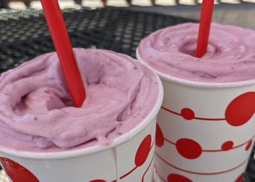 Two cups with purple smoothies topped with whipped cream and red straws, set against a black table.