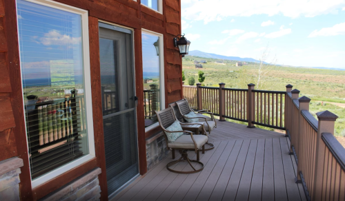 A wooden deck with two chairs overlooks a scenic landscape and mountains under a blue sky.