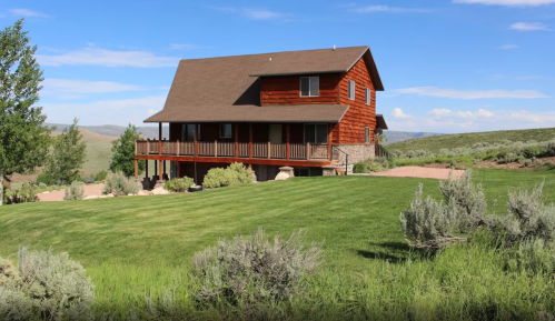 A two-story red wooden house with a porch, surrounded by green grass and shrubs under a blue sky.