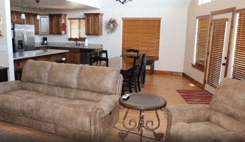 Cozy living area with brown sofas, a small table, and a dining space with wooden furniture and large windows.