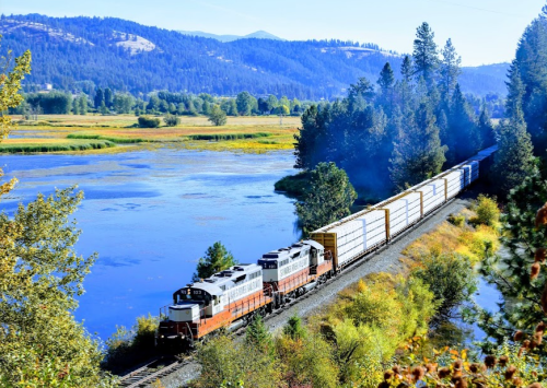 A train travels along a scenic route by a river, surrounded by trees and mountains in the background.
