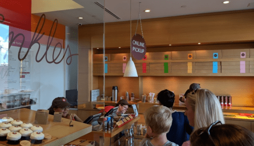 A busy cupcake shop with a counter, customers, and colorful cupcake displays in the background.