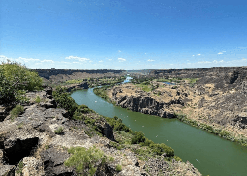 A winding river flows through a rocky canyon under a clear blue sky, surrounded by lush greenery and rugged cliffs.