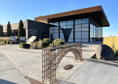 A modern building with large windows, featuring a metal bridge sculpture in the foreground and landscaped surroundings.