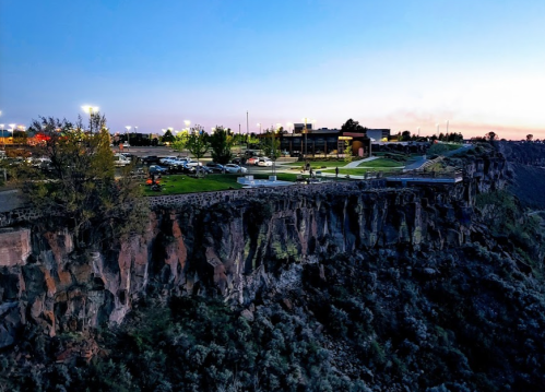 A scenic view of a cliff at dusk, overlooking a parking lot and buildings in the distance, with trees and lights.