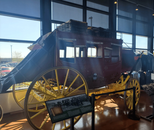 A vintage stagecoach with yellow wheels displayed indoors, surrounded by informational signs and other artifacts.