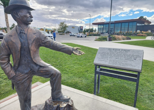 A bronze statue of a man in a suit and hat, gesturing, with a plaque nearby and a modern building in the background.