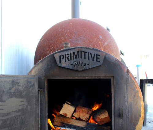 A rustic smoker with a sign reading "Primitive Pits," featuring a fire and stacked wood inside.