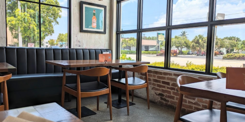 A cozy restaurant interior with wooden tables, chairs, and large windows overlooking a sunny street.