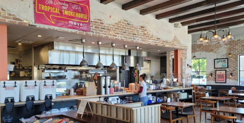 Interior of a restaurant featuring a smokehouse kitchen, wooden tables, and a menu banner hanging on the wall.