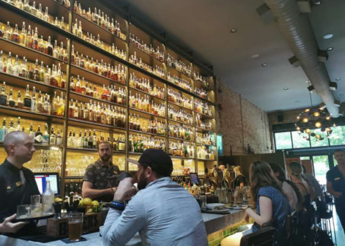 A stylish bar with a wall of liquor bottles, patrons seated at the counter, and bartenders preparing drinks.