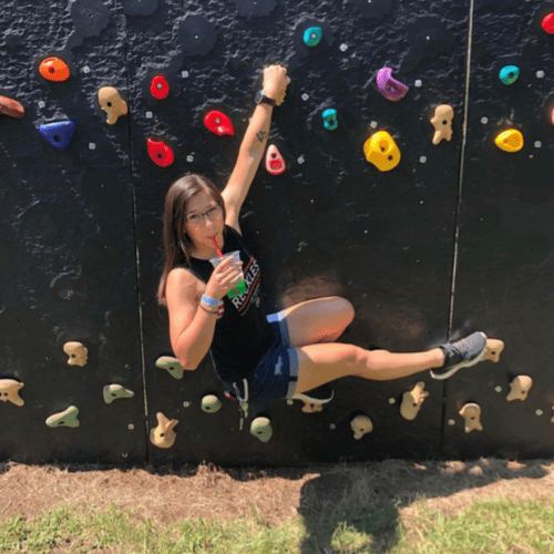 A person climbs a colorful rock wall while holding a drink, wearing a tank top and shorts, enjoying a sunny day.