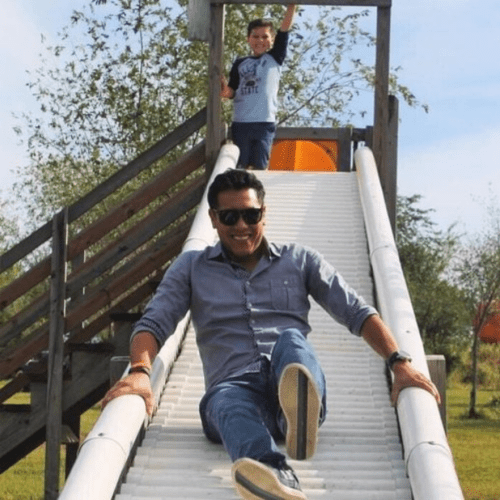 A man in sunglasses slides down a playground slide while a boy stands playfully at the top.