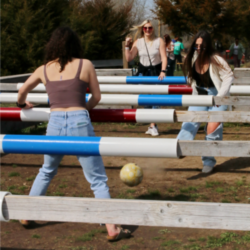 Three young women play a game of human foosball outdoors, with colorful poles and a soccer ball on the ground.