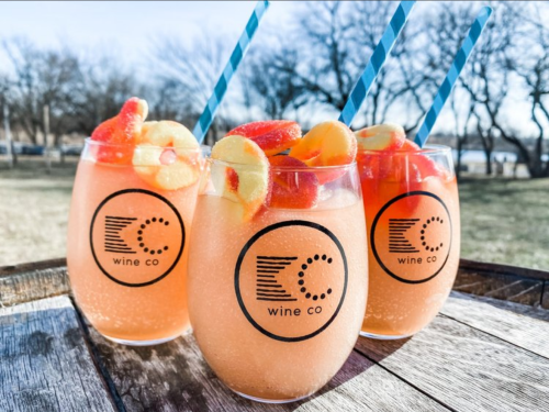 Three glasses of fruity pink drinks with straws, garnished with apple slices and strawberries, on a wooden table outdoors.