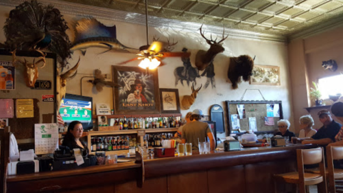 A rustic bar with taxidermy decor, patrons at the counter, and a bartender serving drinks in a cozy atmosphere.