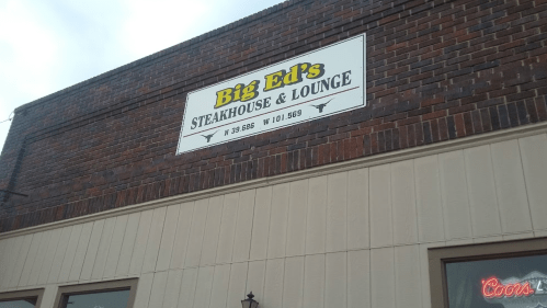 Sign for Big Ed's Steakhouse & Lounge, featuring the restaurant's name and coordinates, against a brick wall.