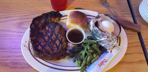 A plate with a grilled steak, green beans, a baked potato, a roll, and a small cup of sauce.