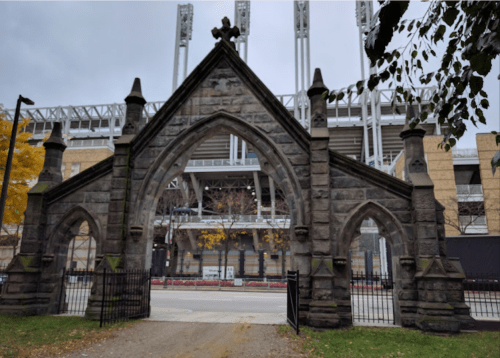 Gothic stone archway with a cross, framed by trees, in front of a modern stadium.