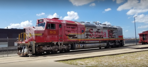 Red and silver train engine with the number 93 parked on a sunny day, surrounded by a fence and blue sky.