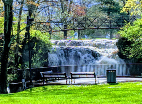 A scenic view of a waterfall surrounded by greenery, with benches and a bridge in the background.