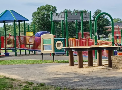 A colorful playground with slides, climbing structures, and a sand area, surrounded by green trees and blue sky.
