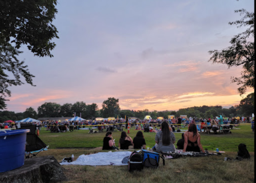 A crowd gathers at a park during sunset, with tents and picnic areas visible in the background.