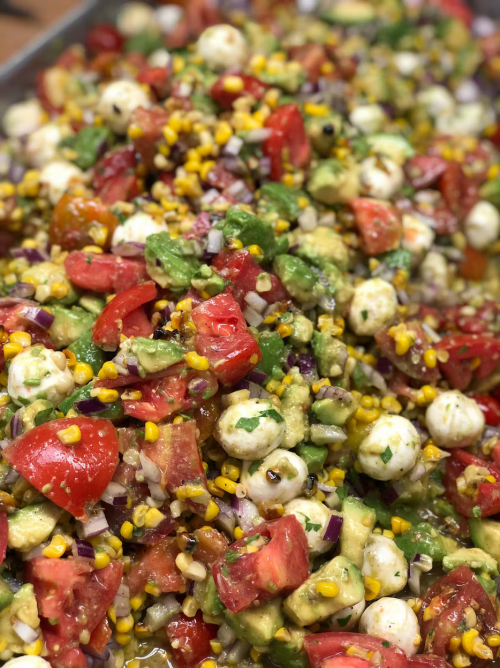 A colorful salad featuring tomatoes, corn, avocado, mozzarella balls, and red onion, mixed in a vibrant dressing.
