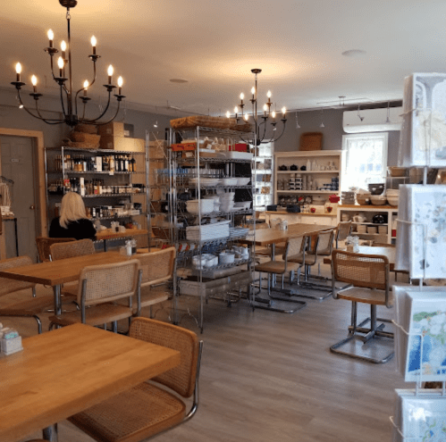 A cozy café interior with wooden tables, shelves of kitchenware, and a chandelier, featuring a person sitting at a table.