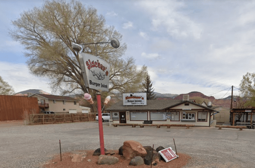 Sign for "Slacker's Burger Joint" with a rustic building and trees in the background, set in a gravel area.