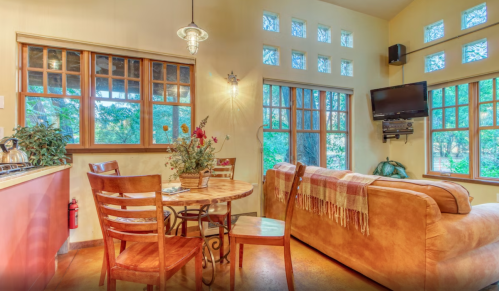 Cozy living space with a sofa, dining table, and large windows letting in natural light. Greenery visible outside.