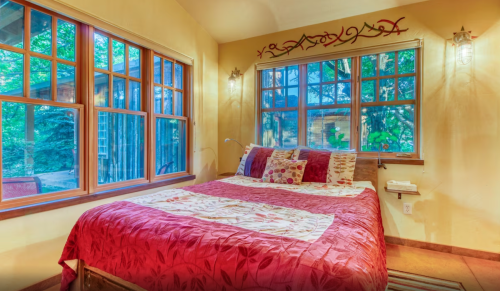 Cozy bedroom with a red quilt, large windows, and warm lighting, surrounded by greenery outside.