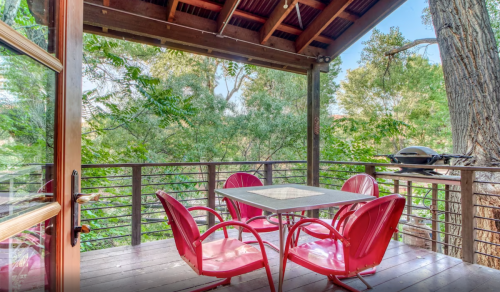 A cozy outdoor deck with a table and red chairs, surrounded by lush green trees.
