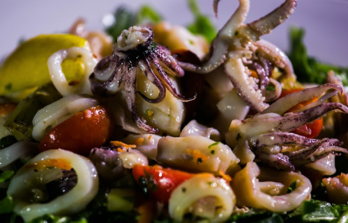 A close-up of a seafood salad featuring squid, octopus, cherry tomatoes, and greens, garnished with lemon.