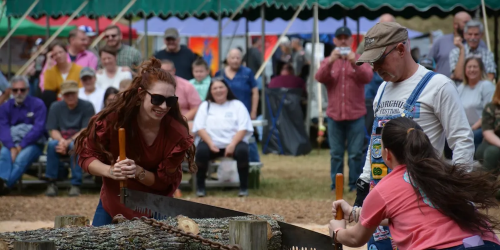 Two participants compete in a log sawing contest, while a crowd watches in the background.