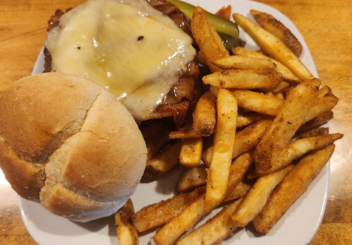 A plate with a cheeseburger, pickles, and a side of golden-brown French fries, alongside a soft bread roll.