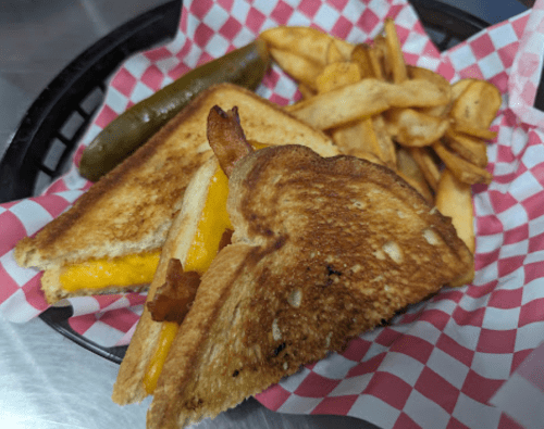 A grilled cheese sandwich with bacon, a pickle, and fries in a basket lined with red and white checkered paper.