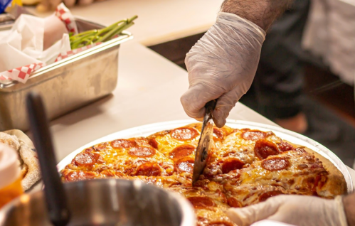A gloved hand slices a pepperoni pizza on a countertop, with kitchen items in the background.