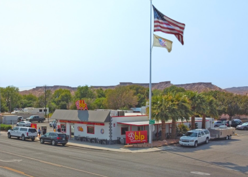 A roadside restaurant with a large American flag, surrounded by palm trees and mountains in the background.