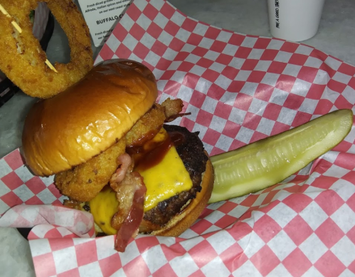 A stacked burger with cheese, bacon, onion rings, and barbecue sauce, served with a pickle on a checkered paper.