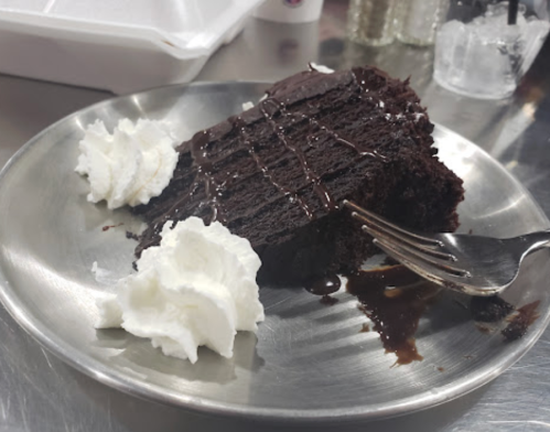 A slice of chocolate cake drizzled with sauce, served on a metal plate with whipped cream on the side.