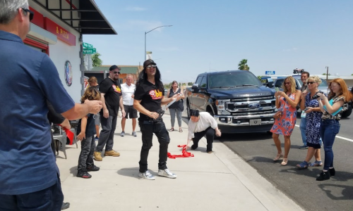 A crowd gathers on the sidewalk, watching a performer in a black outfit and sunglasses, with a truck parked nearby.