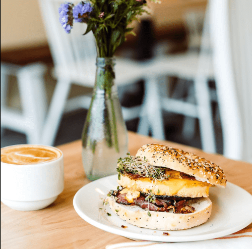 A bagel sandwich with egg and sprouts on a plate, next to a cup of coffee and a vase of flowers on a wooden table.