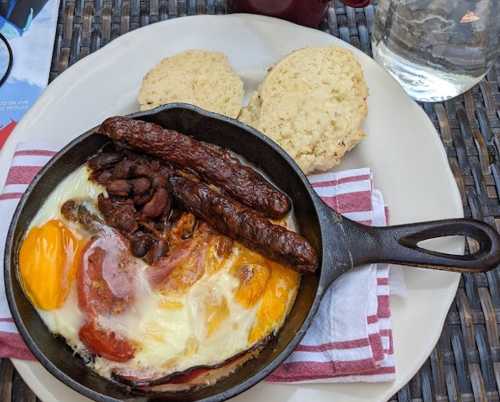 A cast iron skillet with eggs, sausage, bacon, beans, and two biscuits on the side, served on a plate.