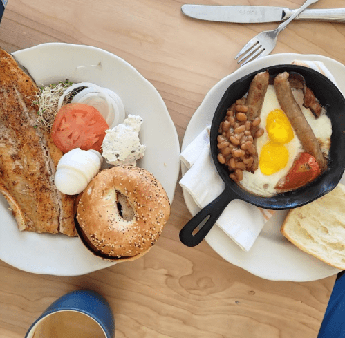 A breakfast spread featuring grilled fish, a bagel, eggs, sausage, baked beans, and sliced tomatoes on a wooden table.
