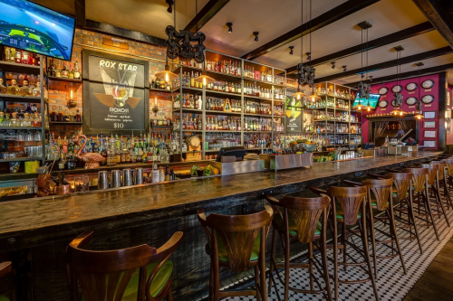 A stylish bar with a long countertop, shelves filled with various bottles, and vibrant decor in a cozy setting.